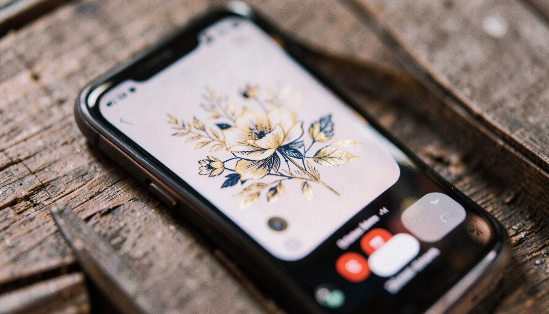 A close-up of a mobile phone screen displaying a hand-drawn floral illustration in soft gold and navy tones, resting on a textured wooden surface